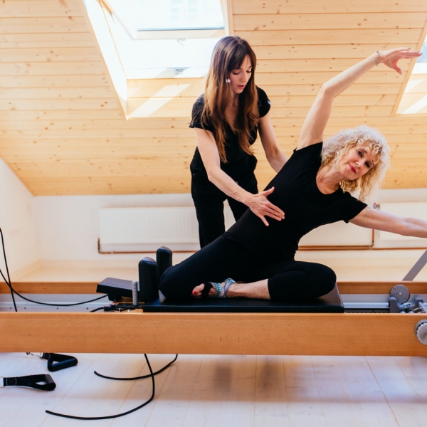 Sporty blond curly senior woman doing pilates exercises in gym with help from female physical therapist or instructor.